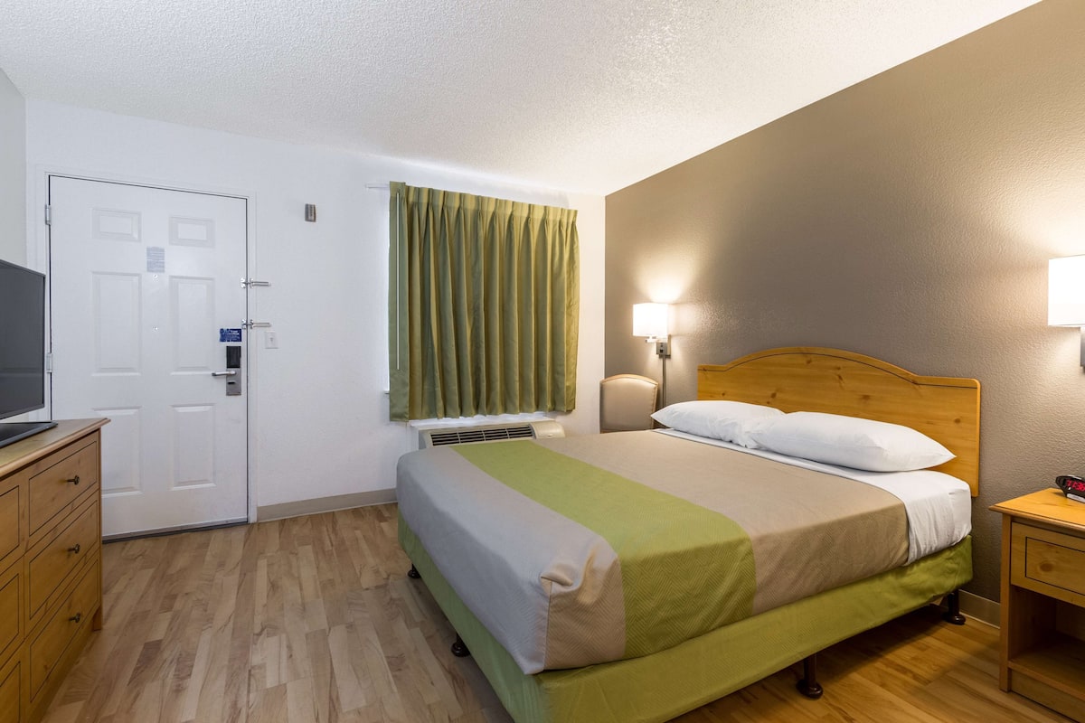 A hotel room with a bed, a desk, a television, a window with curtains, and a wall-mounted lamp.