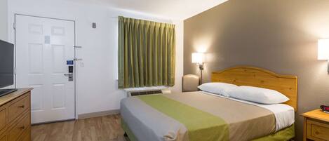 Una habitación de hotel con cama, escritorio, televisión, ventana con cortinas y lámpara en la pared.