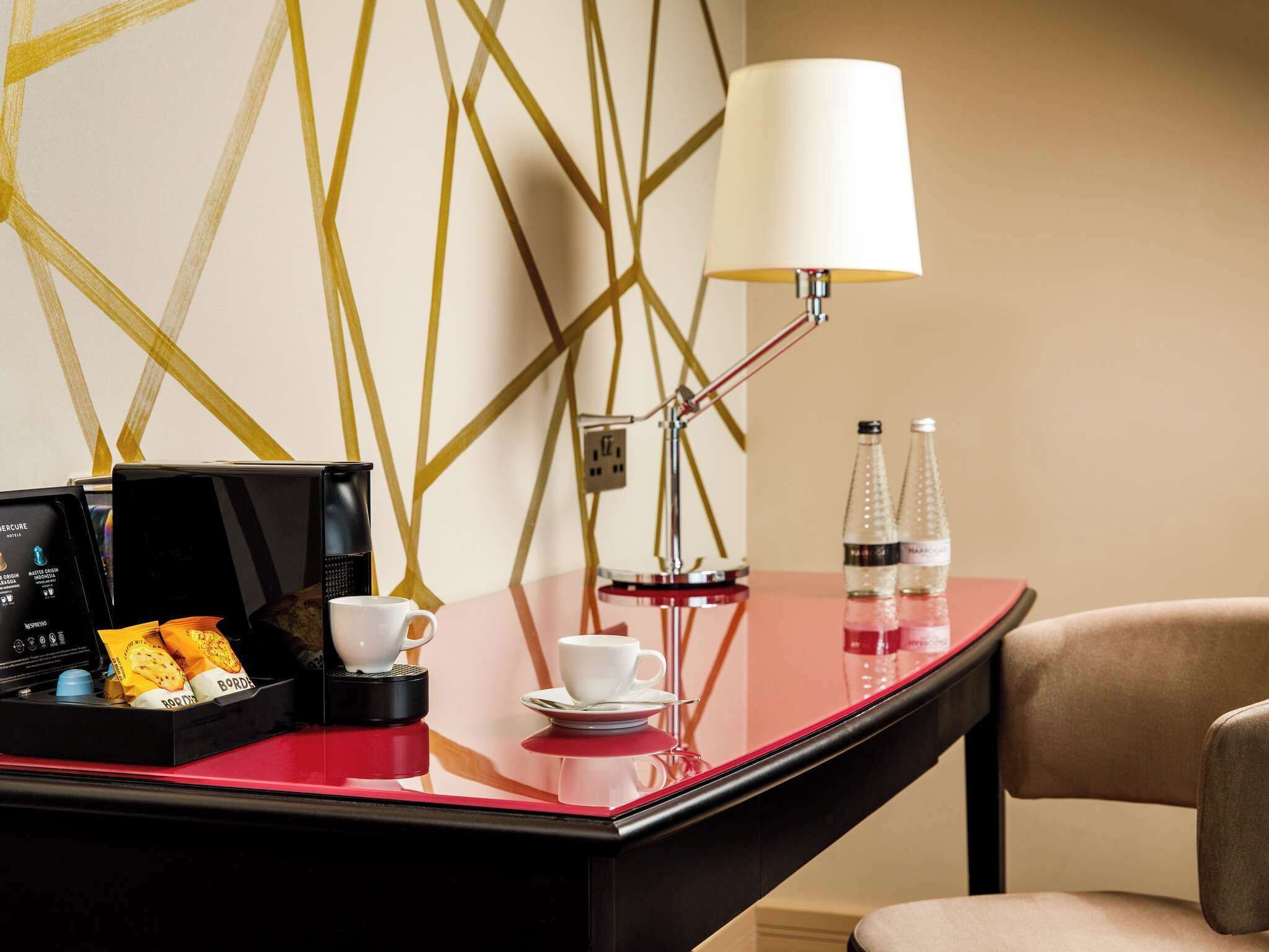 A hotel room with a red table, a coffee machine, a cup, a saucer, and two bottles of water.
