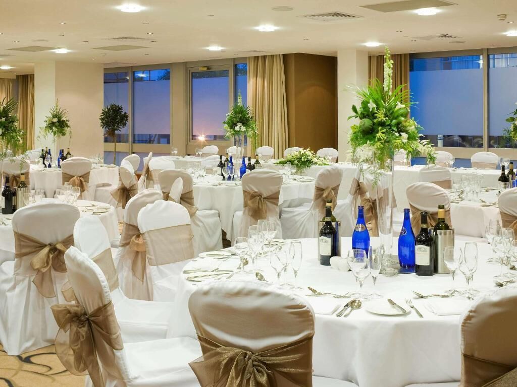 Une salle de banquet avec des tables rondes dressées pour un événement formel, agrémentée de nappes blanches, de verres et de compositions florales.