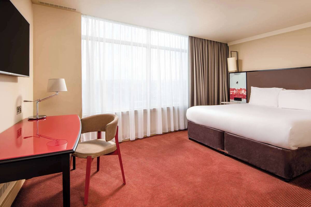 Hotellihuone, jossa on sänky, työpöytä, tuoli ja suuri ikkuna, jossa on verhot.