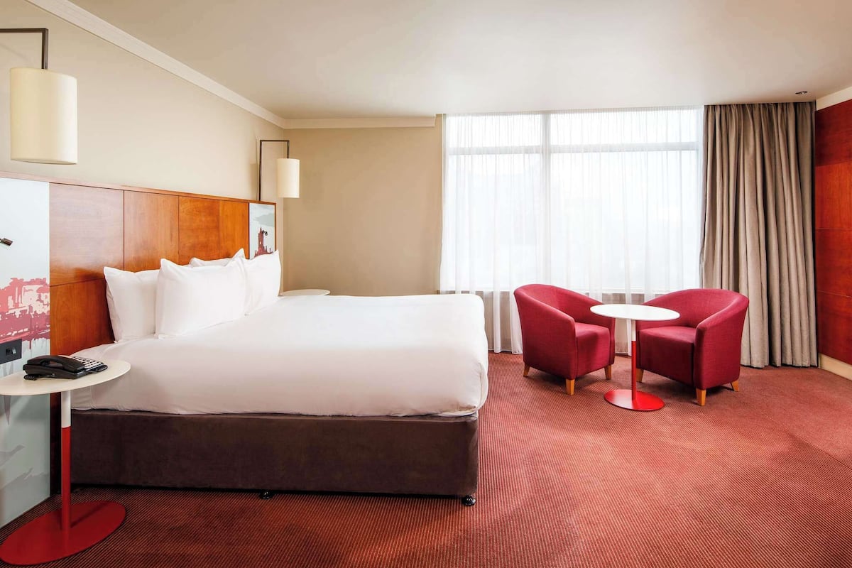 Hotellihuone, jossa on suuri sänky, kirjoituspöytä puhelimineen, kaksi punaista nojatuolia, pieni pyöreä pöytä ja suuri ikkuna, jossa on verhot.
