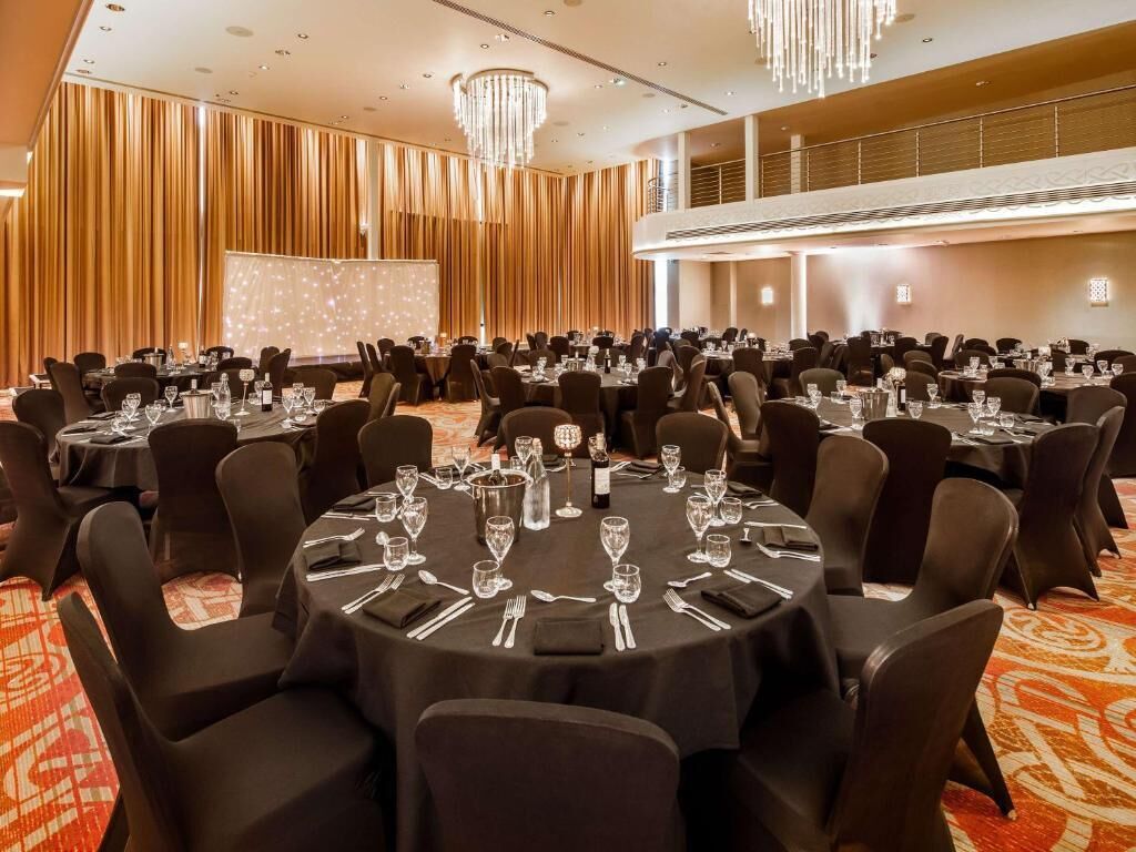Ein Festsaal mit runden Tischen zum Essen, eine Bühne mit einer Projektionsleinwand und ein Balkon mit Blick auf den Raum.