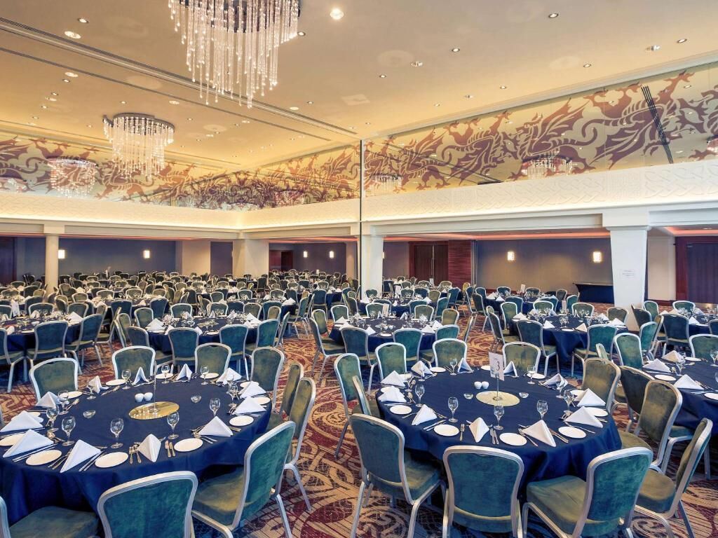 Une salle de banquet avec des tables rondes dressées pour le repas, un plafond haut orné de lustres et une fresque décorative sur le mur.