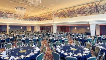 A banquet hall with round tables set for dining, a high ceiling with chandeliers, and a decorative mural on the wall.