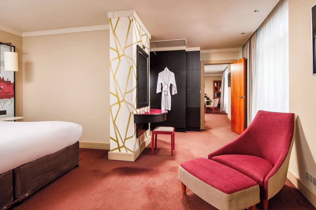 Hotellihuone, jossa on sänky, punainen nojatuoli, pieni pöytä, televisio ja vaatekaappi.