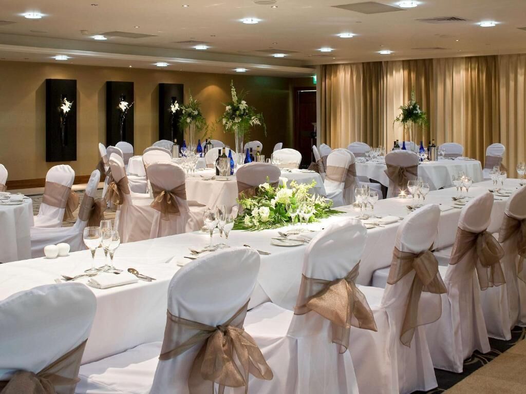 Un salón de banquetes con mesas redondas, manteles blancos y sillas con cintas doradas.