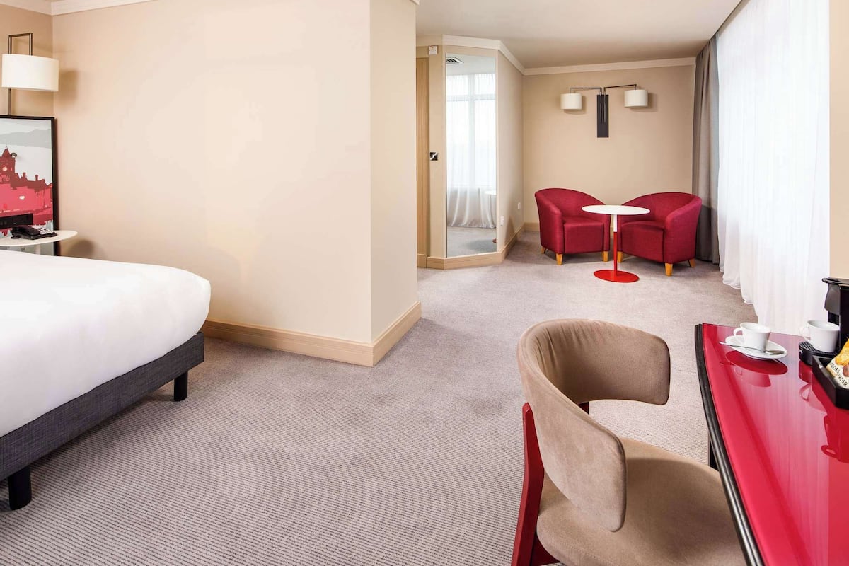 Hotellihuone, jossa on sänky, työpöytä tuolilla, punainen nojatuoli ja pieni pöytä.