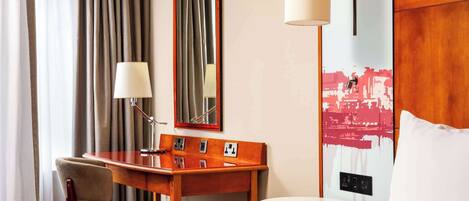 Habitación de hotel con una cama, un escritorio con una silla, una lámpara, un espejo y un cuadro en la pared.