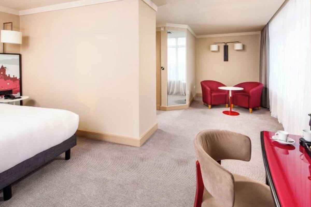 Ett hotellrum med en säng, ett skrivbord med en stol, ett litet bord med två stolar och en dörr som leder till ett annat rum.