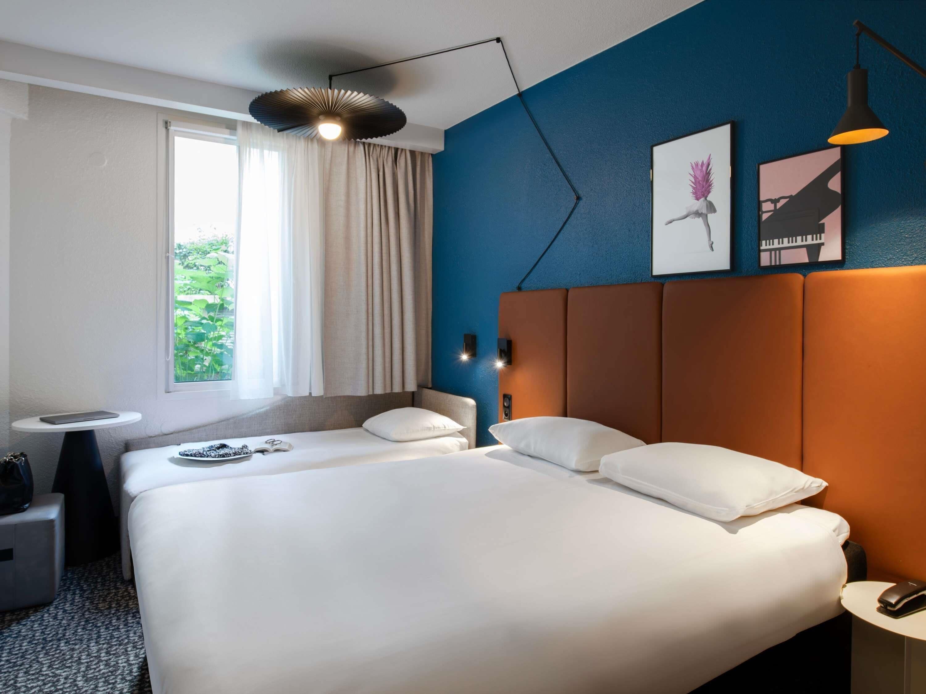 A modern hotel room with a large bed, a small bedside table, a nightstand, and a window with curtains.