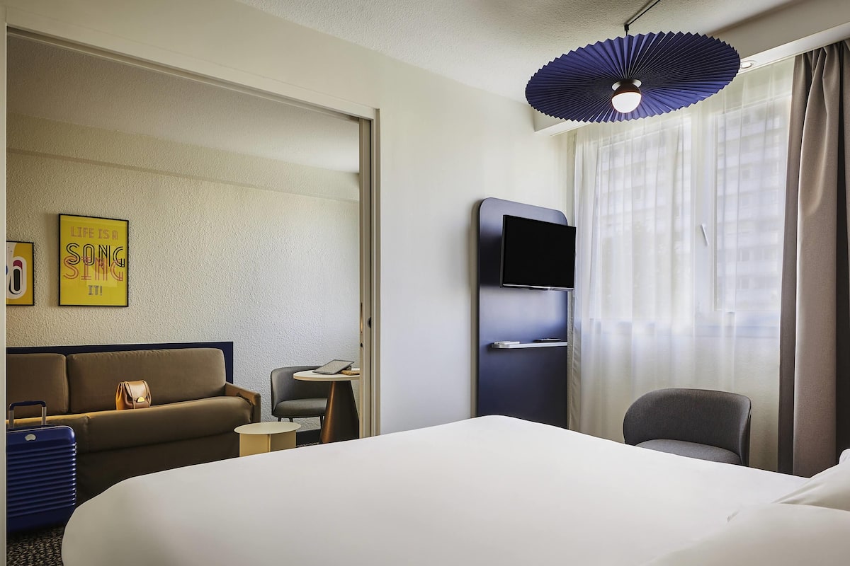 Habitación de hotel con una cama grande, un sofá, un escritorio con una computadora portátil, un televisor y una ventana con cortinas.