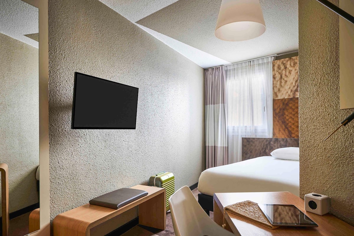Una habitación de hotel moderna con una televisión de pantalla plana montada en la pared, un escritorio de madera con una computadora portátil, una mesita de noche con una tableta, una cama con un almohadón blanco y una ventana con cortinas de gasa.