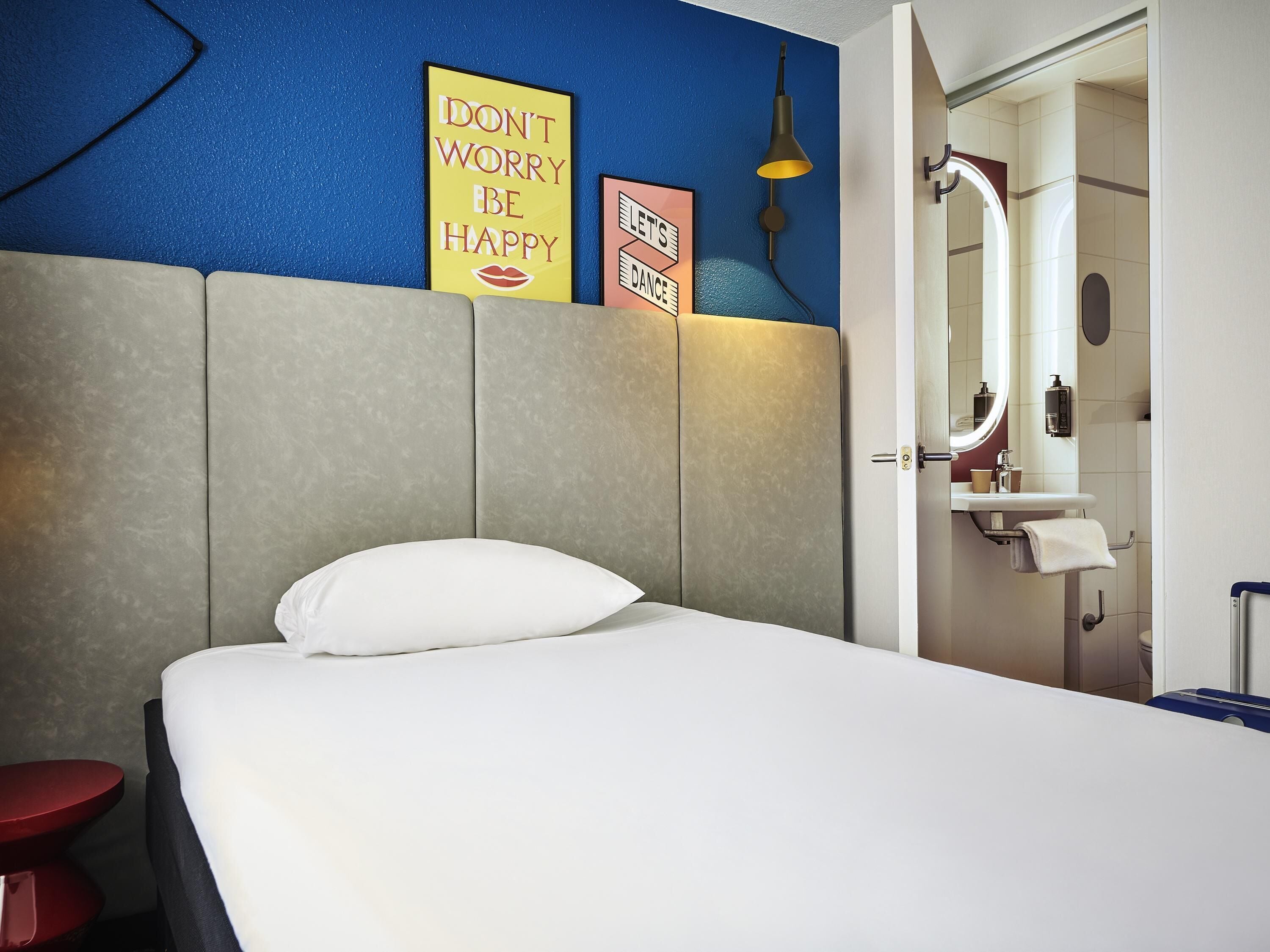 Habitación de hotel con cama, un taburete rojo, un baño con lavabo y espejo, y decoración mural motivacional.