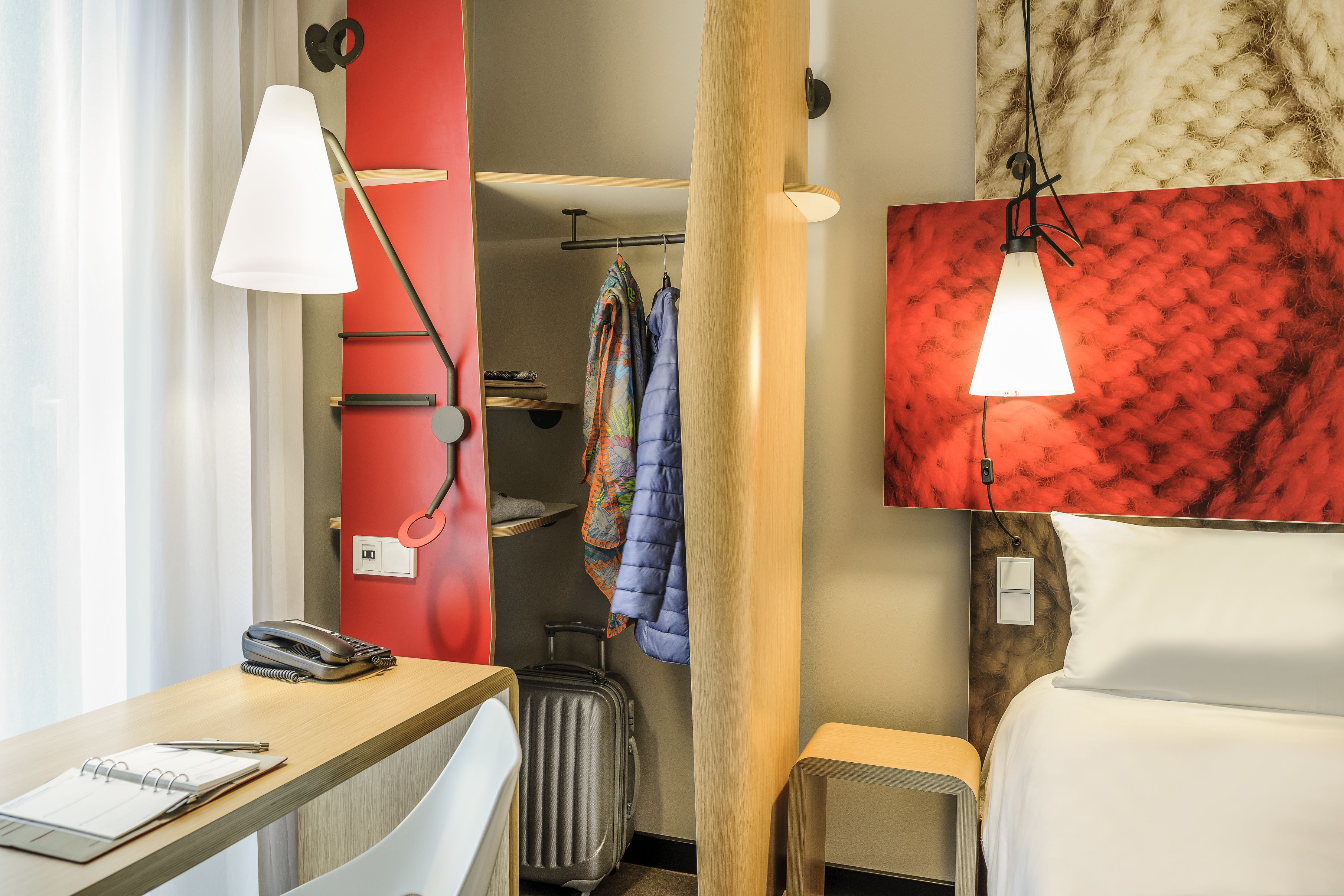 A hotel room with a desk, a telephone, a notebook, a suitcase, a bed, and a wardrobe.