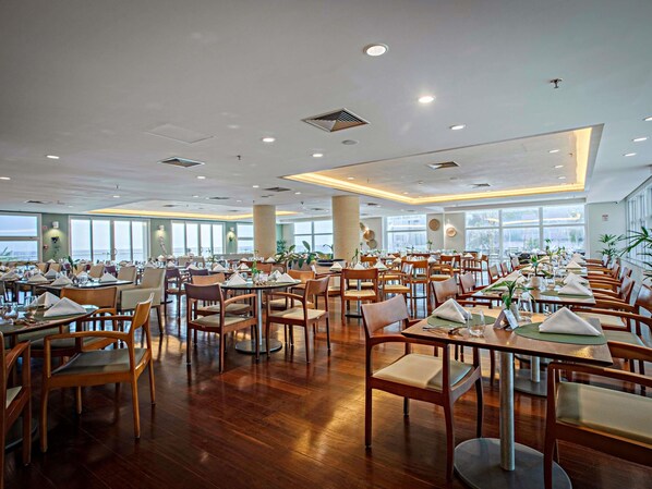 Un vaste espace dédié à la salle à manger, avec de nombreuses tables et chaises, de grandes fenêtres offrant une vue dégagée et un design moderne au niveau du plafond.