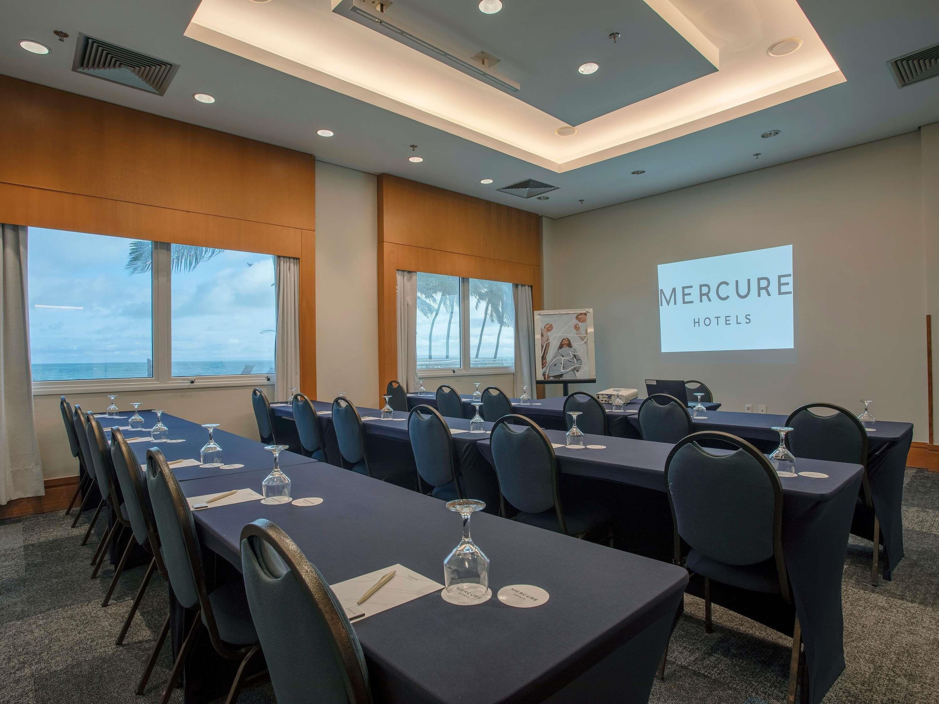 Uma sala de conferência com mesas, cadeiras e uma tela de projeção exibindo 'Mercure Hotels'.
