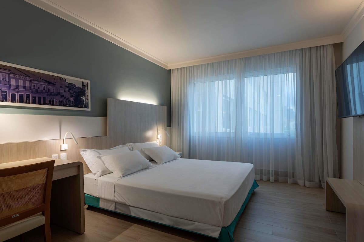 Ein Hotelzimmer mit einem Bett, Nachttischen, einem Schreibtisch, einem Sessel, einem Bild an der Wand und einem Fenster mit Vorhängen.