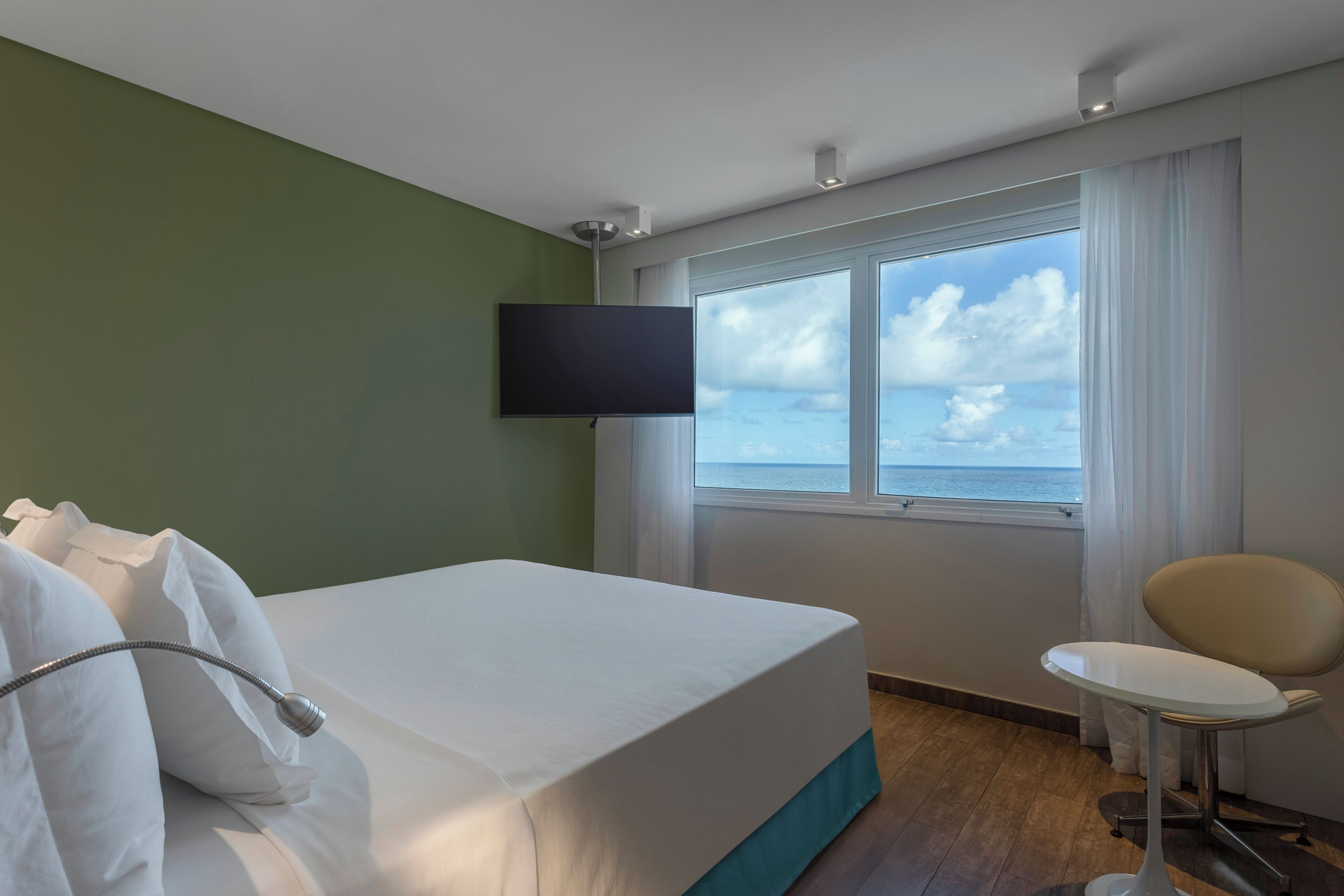 Habitación de hotel con cama, televisión montada en la pared, ventana con vista al mar y una silla.