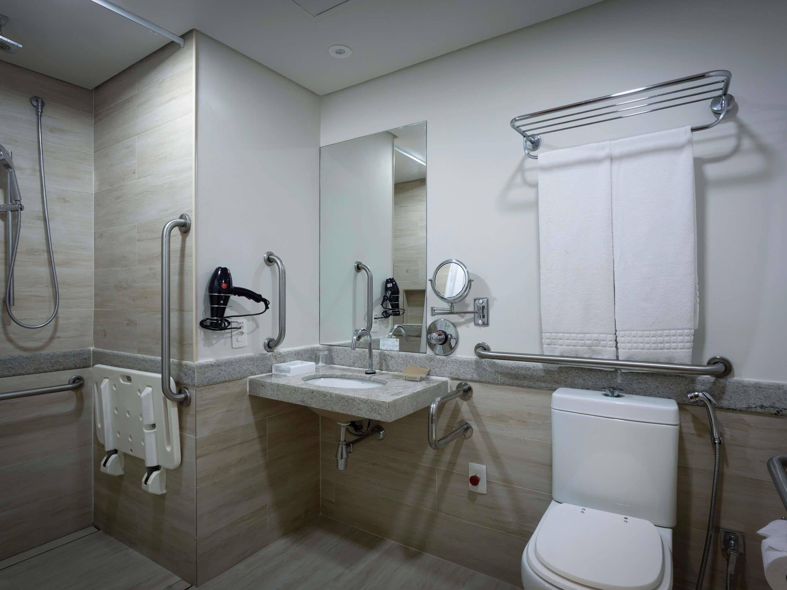 Une salle de bain moderne équipée d’un lavabo, de toilettes et d’une douche. Elle comprend également des barres d’appui et un sèche-cheveux.