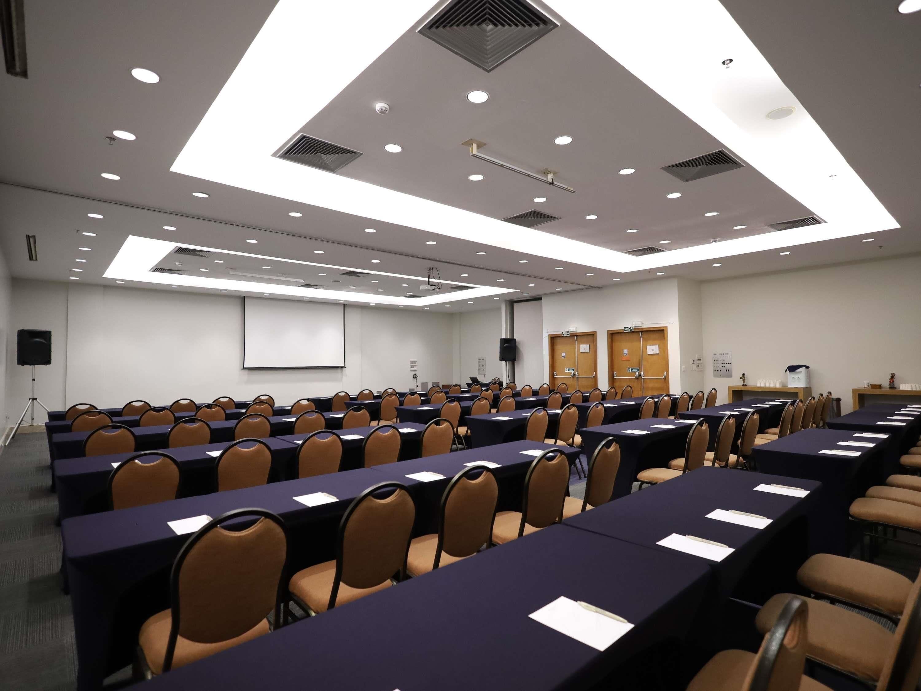 Un salón de conferencias con filas de mesas y sillas, una pantalla de proyector y un orador.