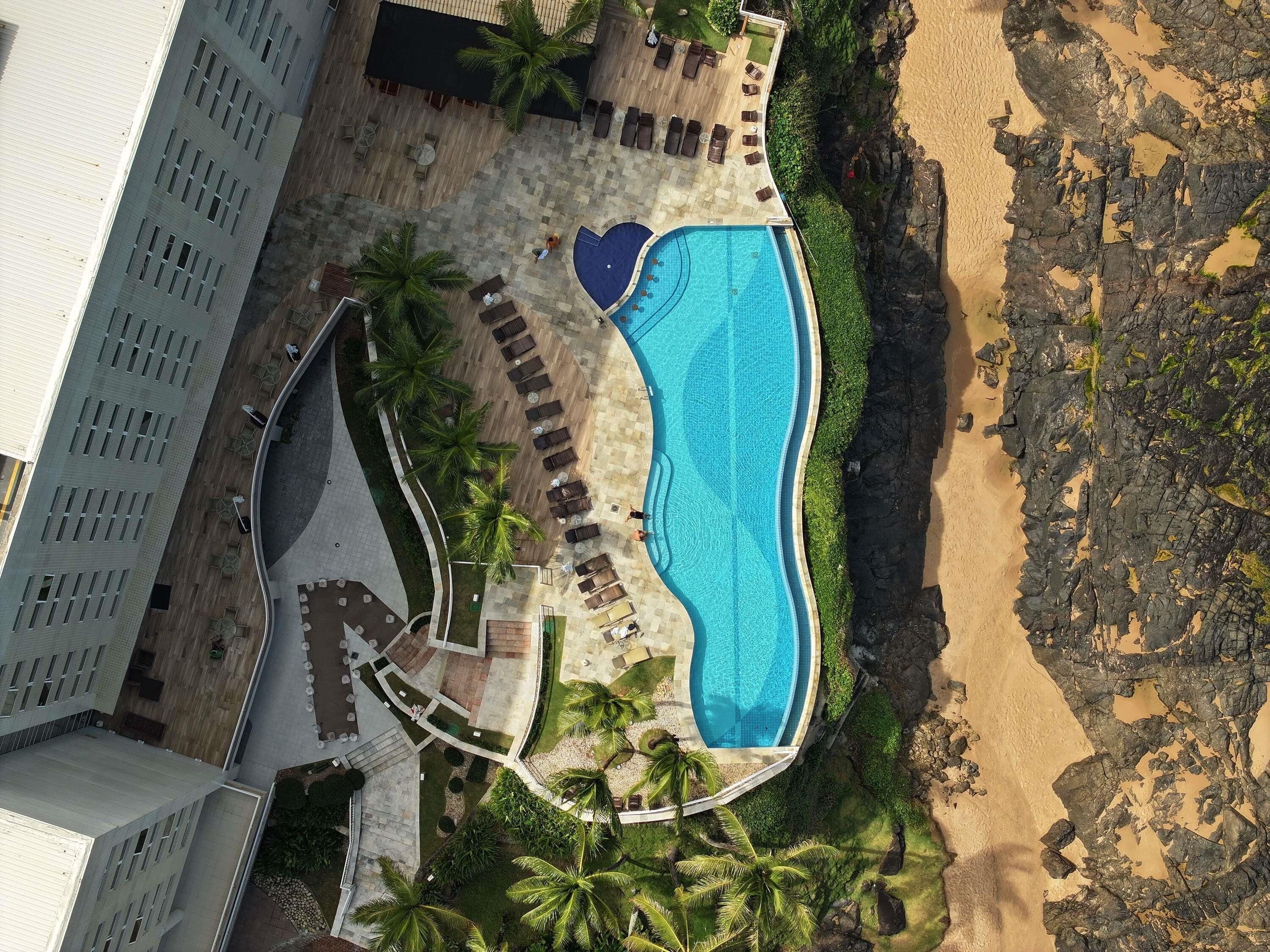 An aerial view of a resort with a swimming pool, lounge areas, and a beach.