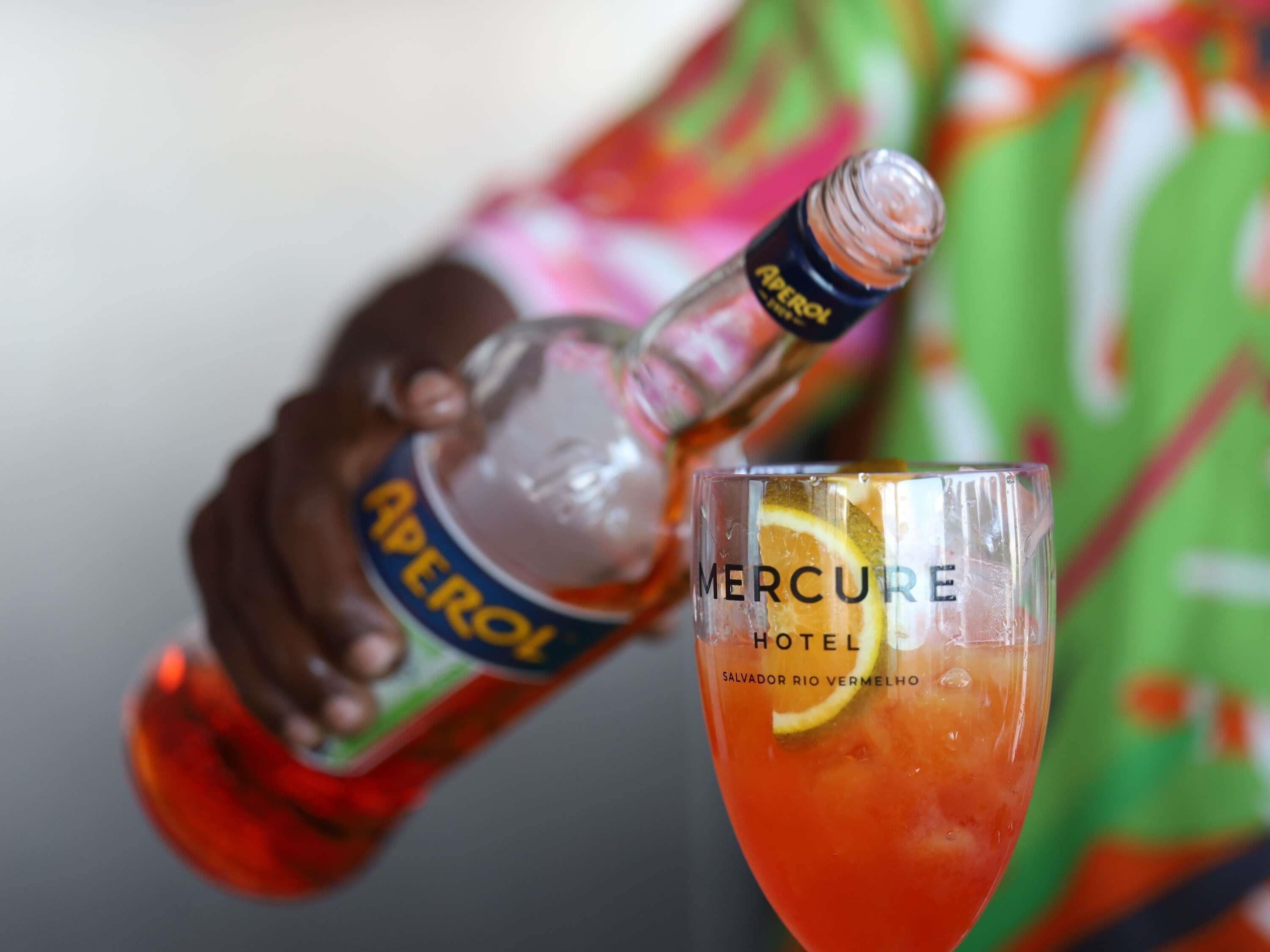 Une bouteille d’Aperol est versée dans un verre, dans lequel on a mis une tranche de citron. Le verre porte les inscriptions « Mercure Hotel » et « Salvador Rio Vermelho ».