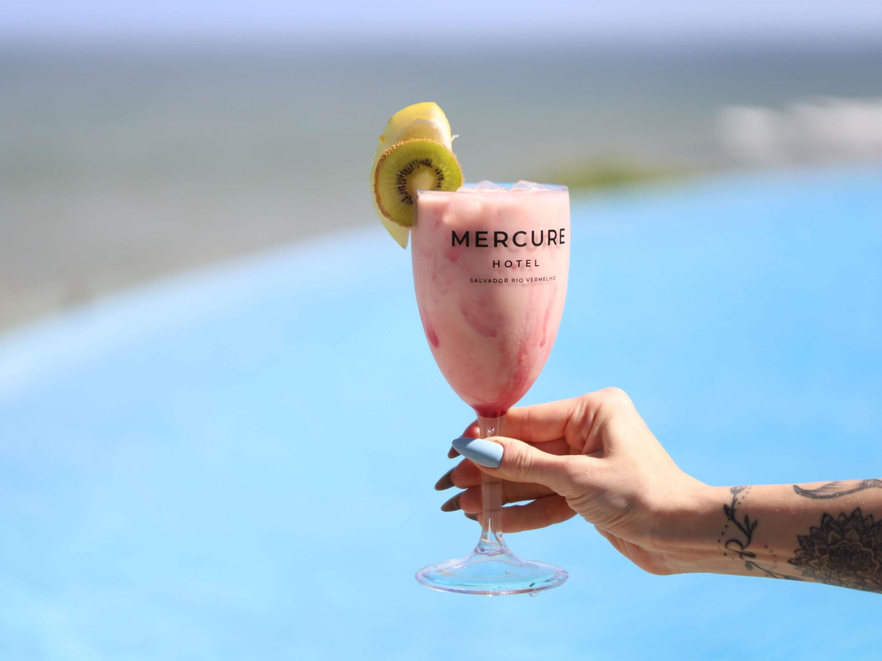 Una mano sostiene un vaso de cóctel rosa con una rodaja de kiwi, con la etiqueta 'Mercure Hotel' en Salvador, Río Vermelho.