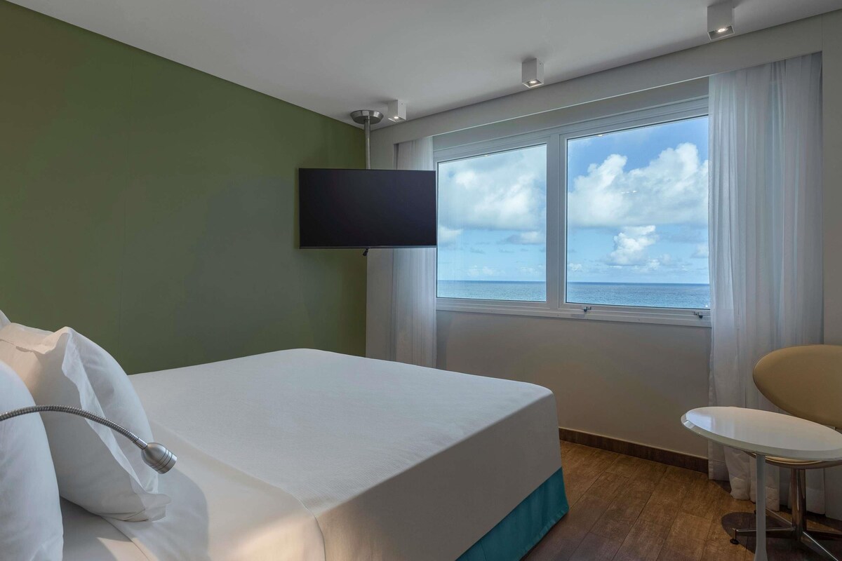 Ein Hotelzimmer mit Bett, Wandfernseher, Fenster mit Meerblick und einem kleinen Tisch.