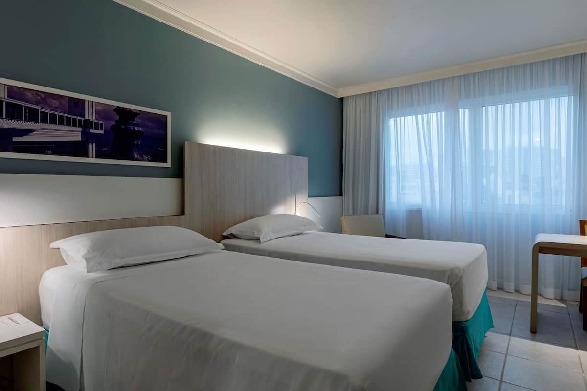 Ein Hotelzimmer mit zwei Betten, einem Schreibtisch, einem Stuhl, einem Bild an der Wand und einem Fenster mit Vorhängen.