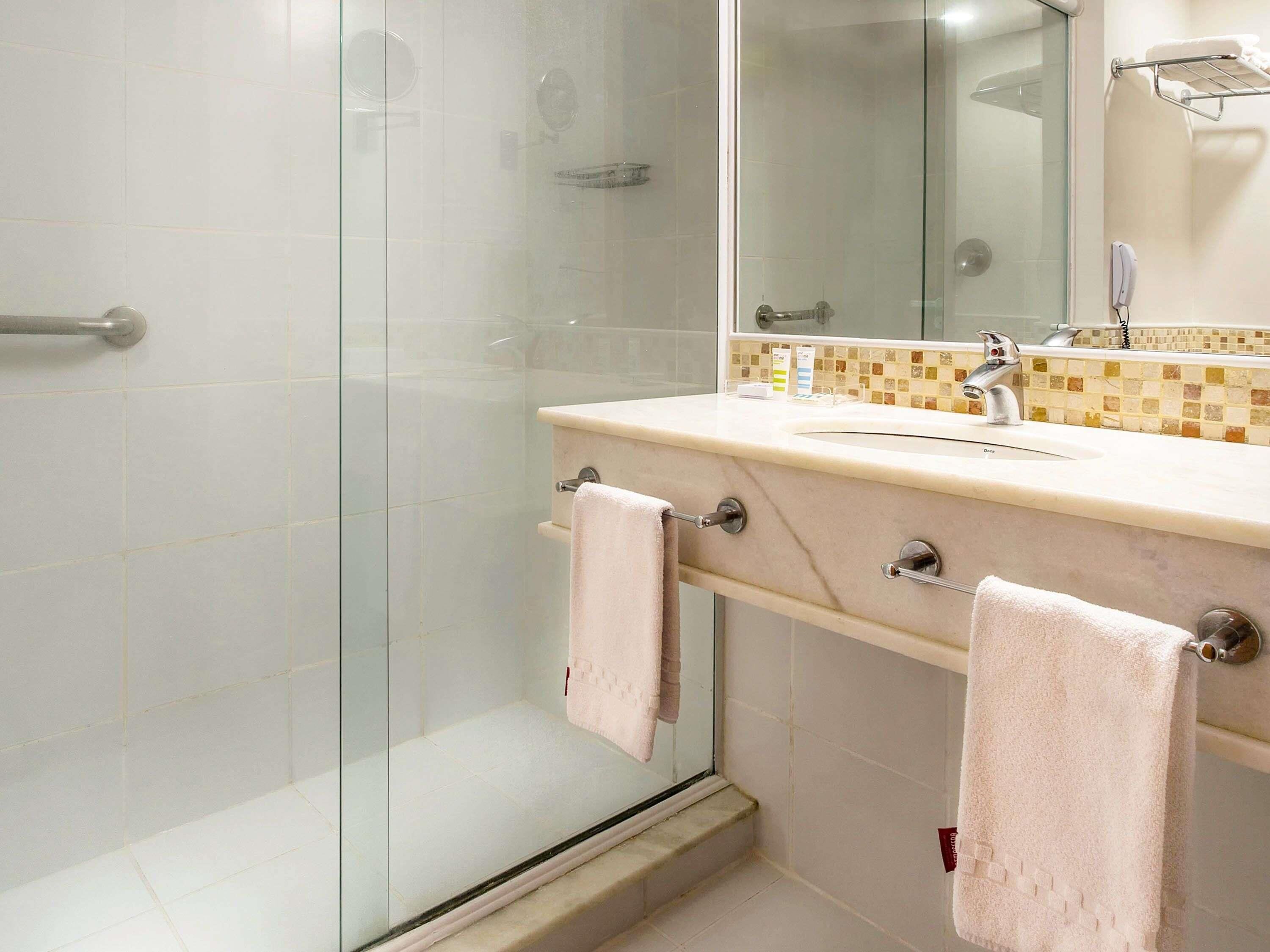A modern bathroom with a glass shower enclosure, a beige tiled wall, a sink with a mirror, and two towels hanging on towel bars.