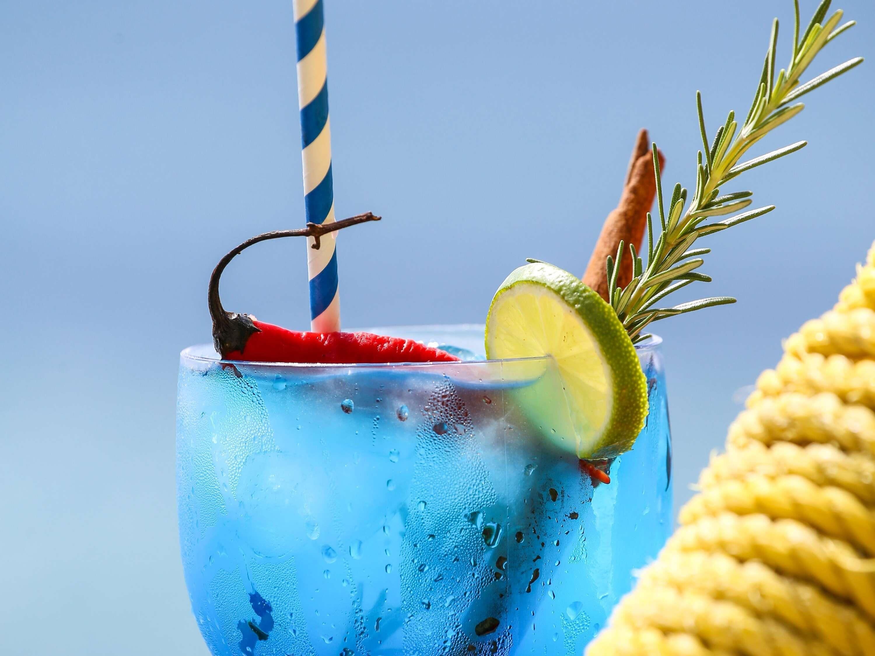 A glass with a blue beverage, a red chili pepper, a lime wedge, and a sprig of rosemary.
