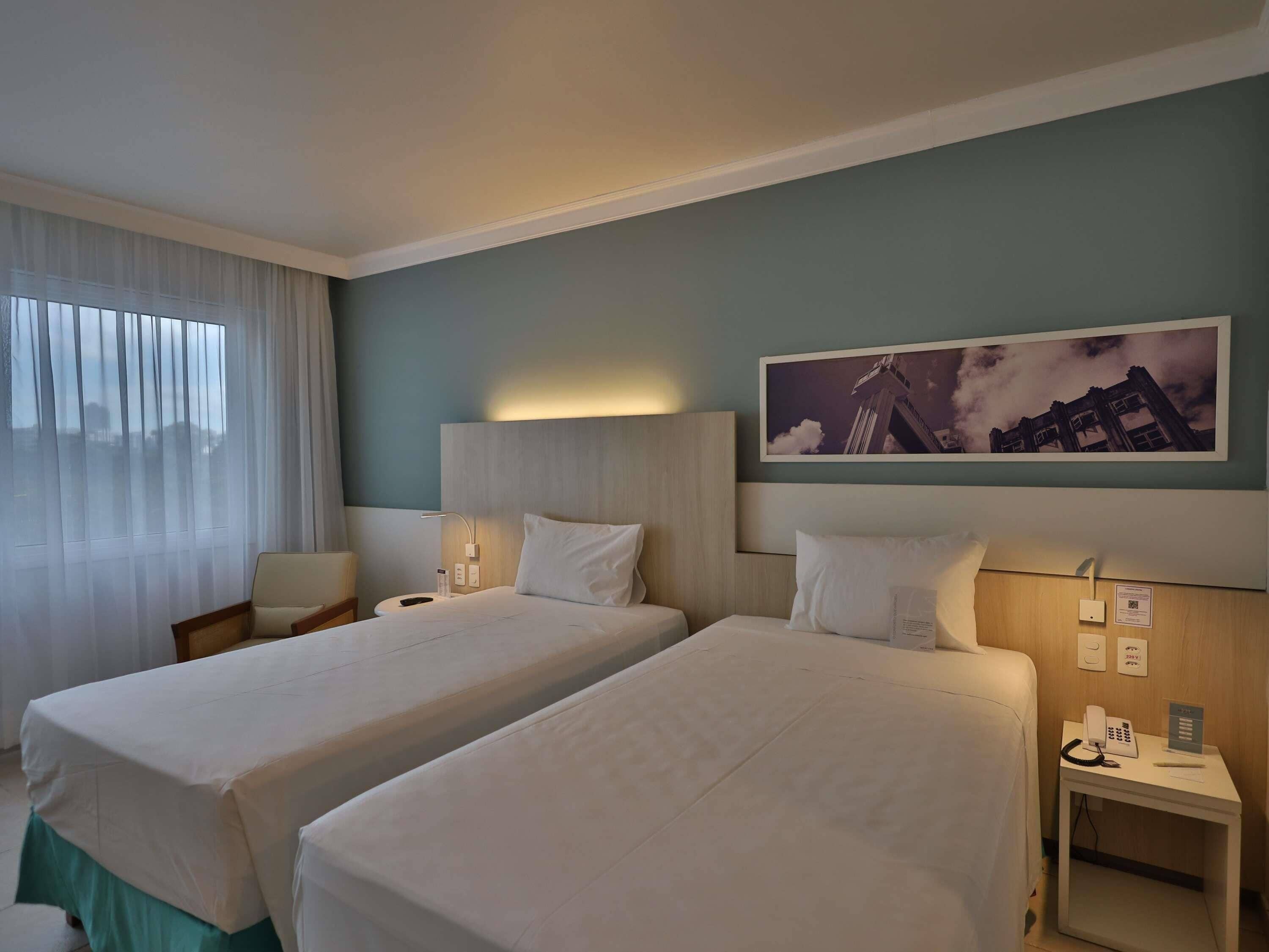 Habitación de hotel con dos camas, una mesita de noche con un teléfono y un cartel, una ventana con cortinas y un cuadro en la pared.
