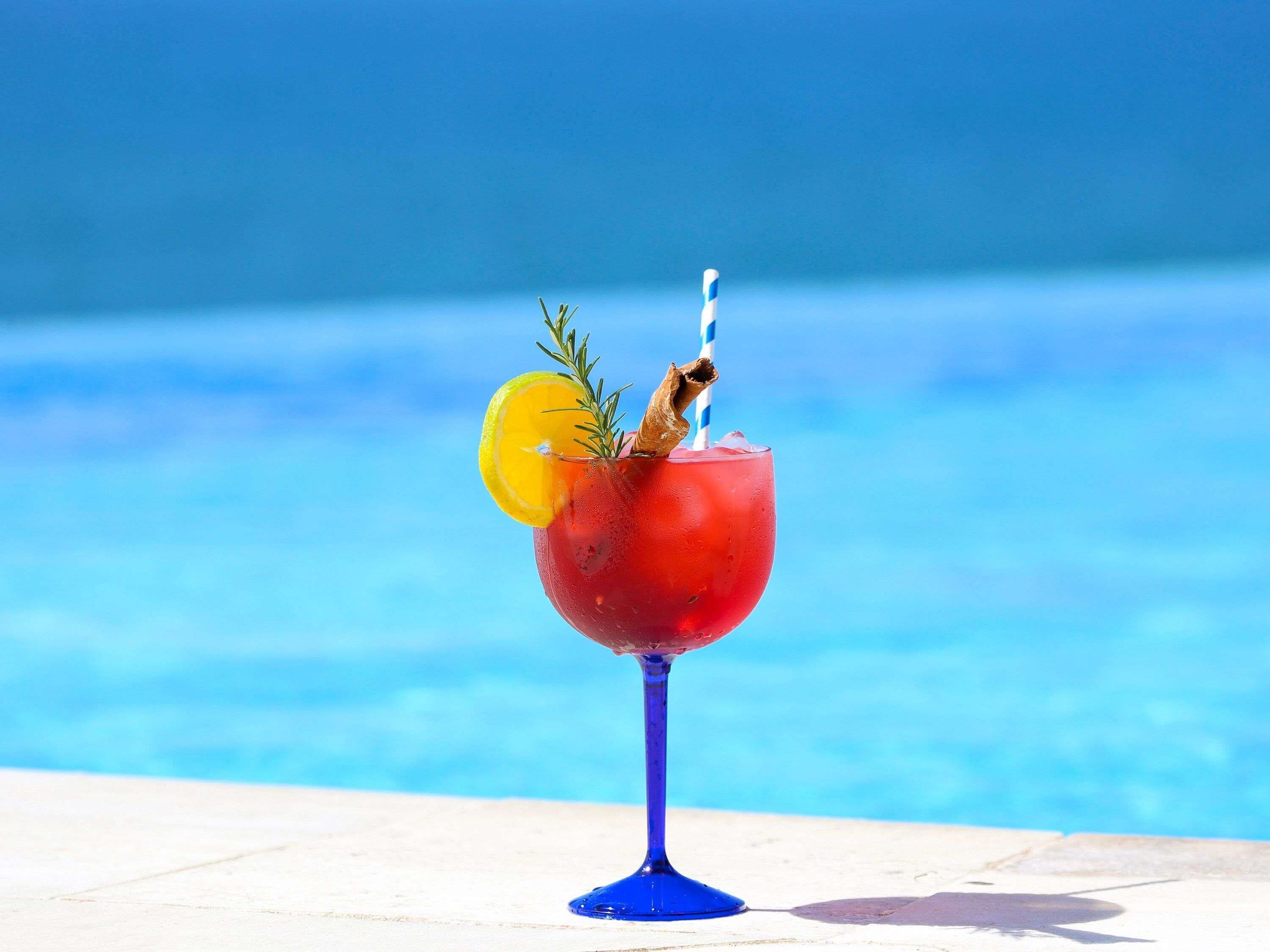 A cocktail with a lemon slice and rosemary garnish, served in a blue glass, by a poolside.