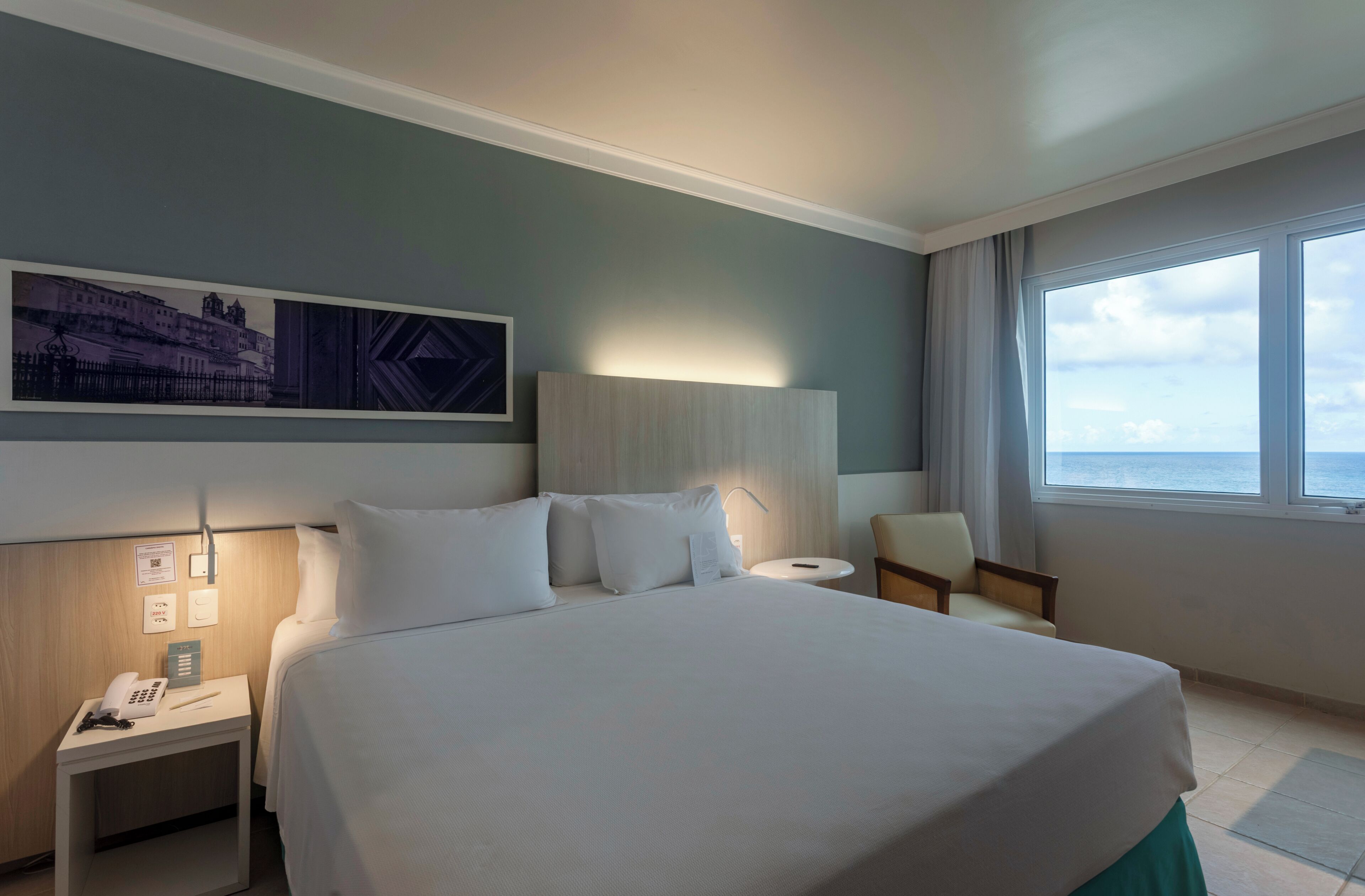 Una habitación de hotel con cama, mesita de noche, una silla, ventana con vistas y una obra de arte colgada en la pared.