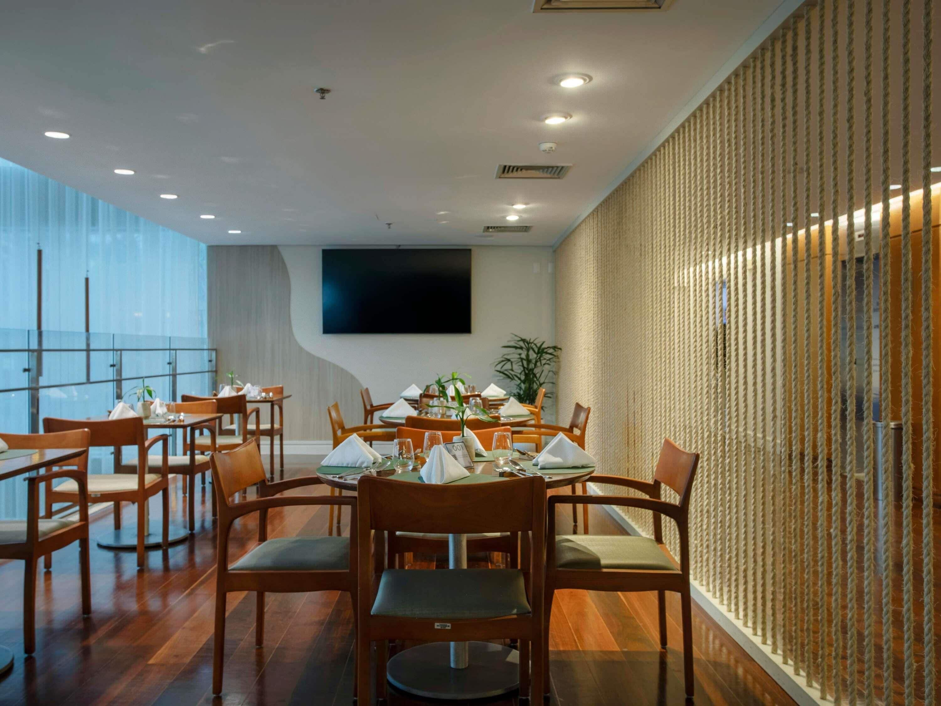 A modern dining area with round tables and wooden chairs, a large flat-screen TV on the wall, and a rope-textured wall on one side.