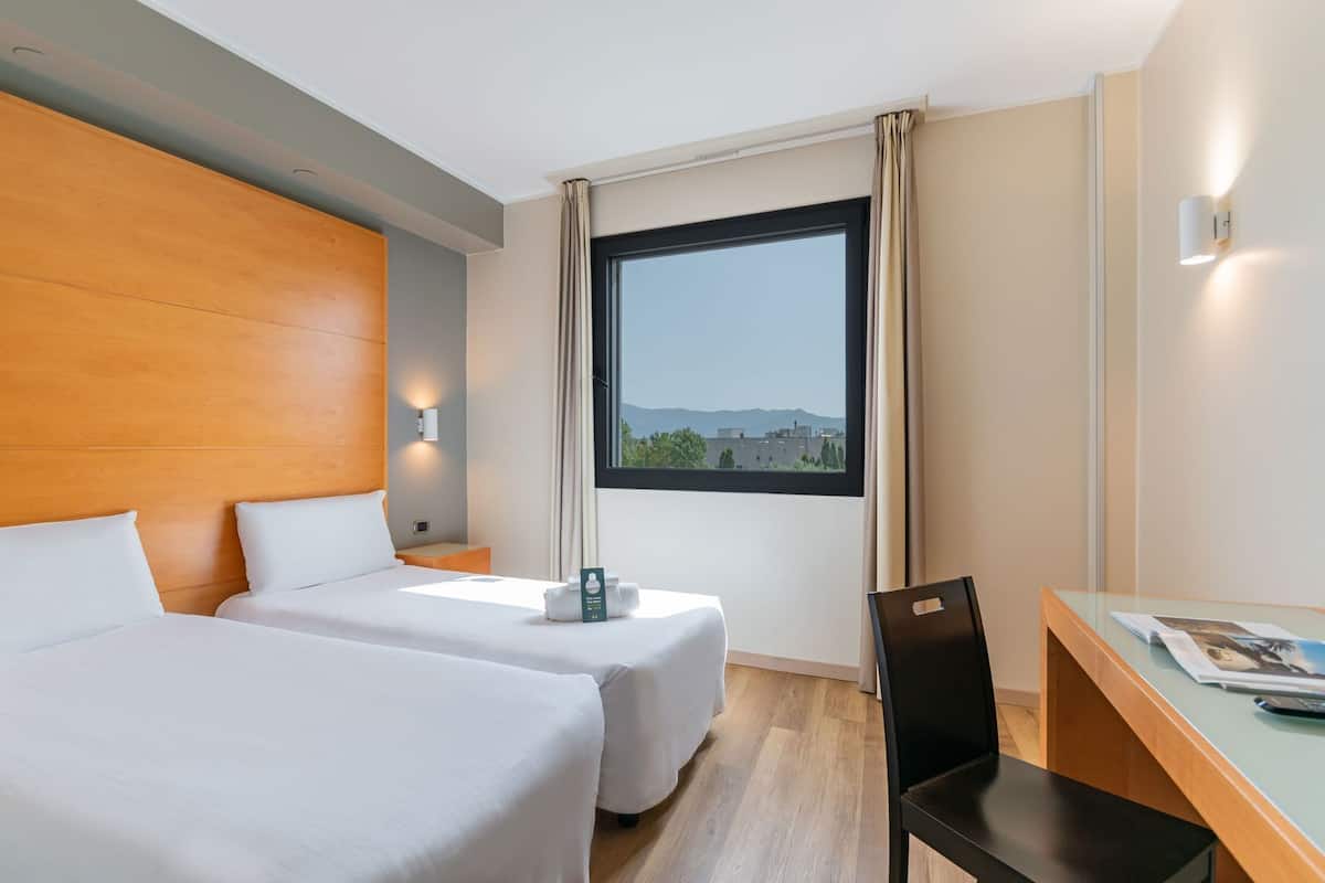 A hotel room with two single beds, a desk with a chair, a window with a view of buildings, and a wall-mounted light.