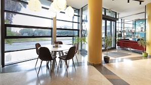A spacious lobby with a large table and chairs, large windows, and potted plants. - B&B HOTEL Pisa Airport (Pisa)