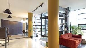 A modern lobby with a red sofa, large pillars, a reception desk, and a glass door leading to the outside. - B&B HOTEL Pisa Airport (Pisa)