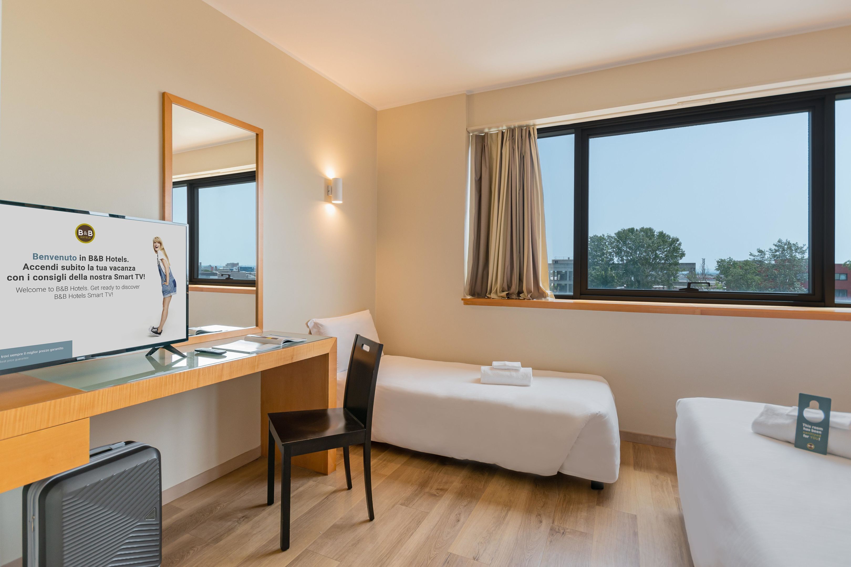 A hotel room with a desk, a chair, a bed, a small bedside table, a TV, and a window with curtains.