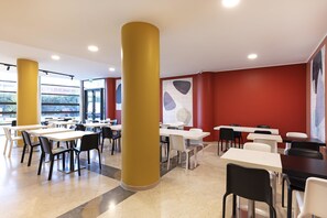 A cafeteria with white tables and chairs, yellow pillars, and a red wall with abstract art. - B&B HOTEL Pisa Airport (Pisa)