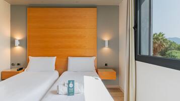 Habitación de hotel con dos camas, cabecero de madera, lámparas de noche, una mesita y una ventana con vistas a zonas verdes.