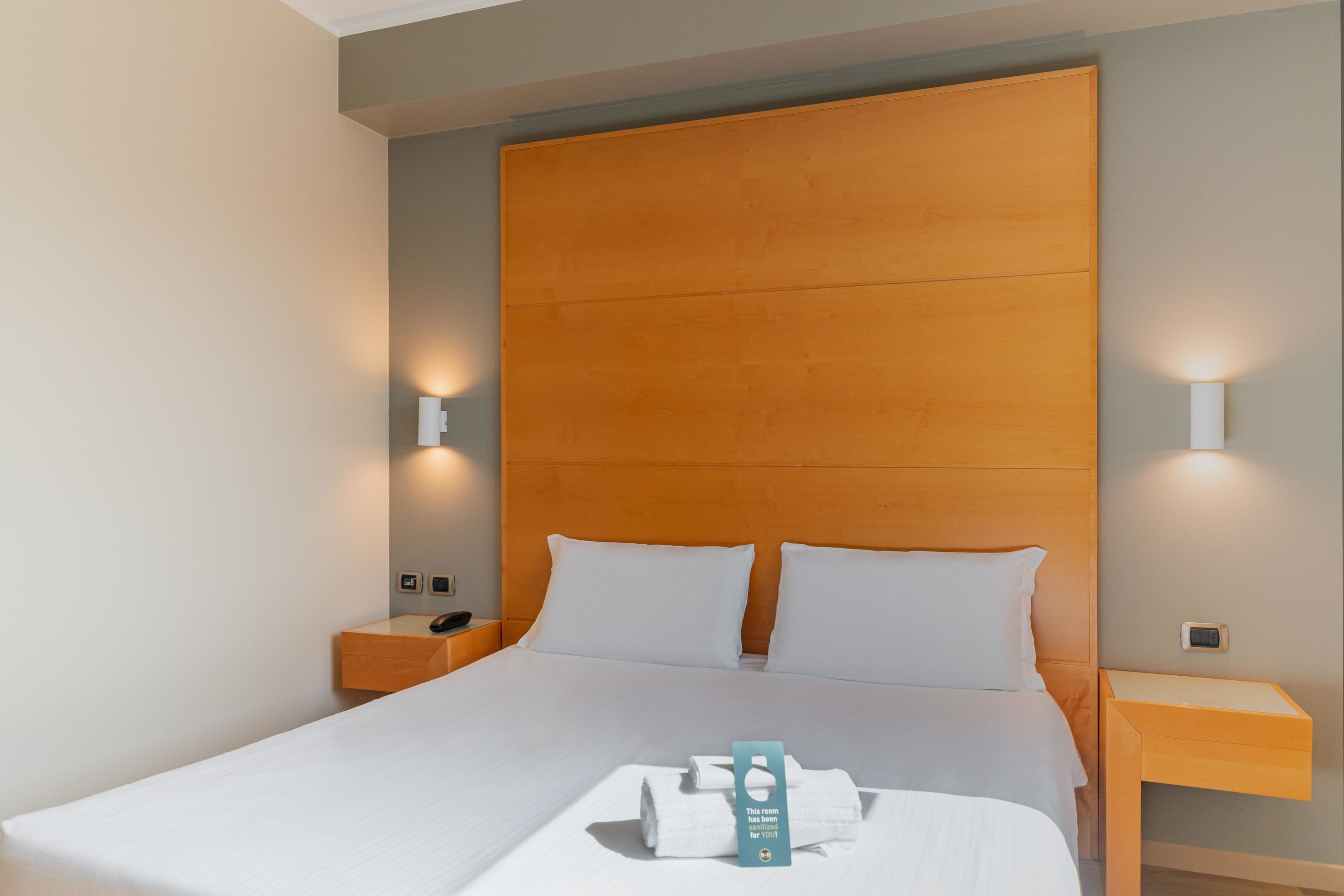 Habitación de hotel con cabecera de madera, ropa de cama blanca y dos lámparas de noche.