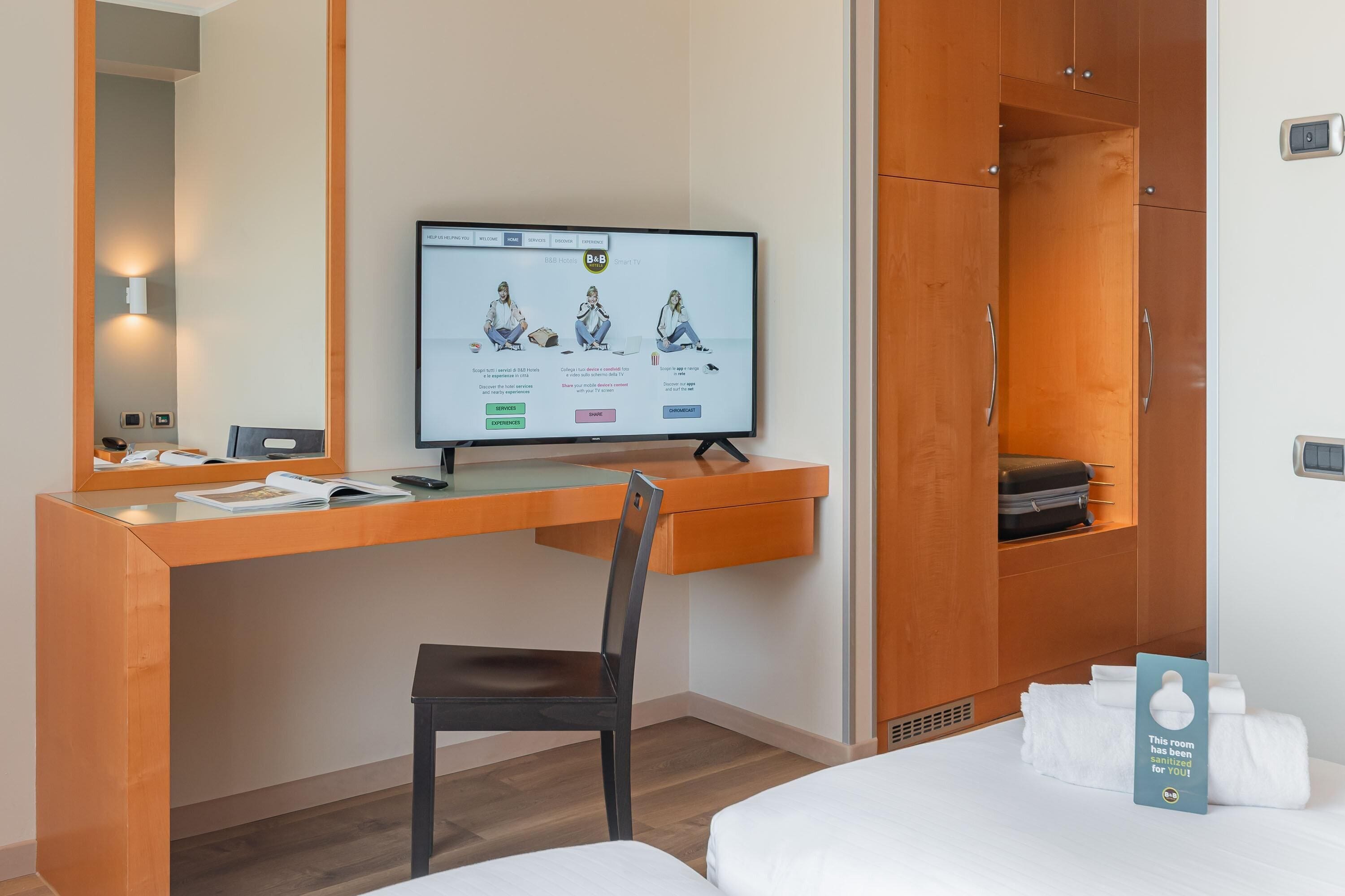 A hotel room with a wooden desk, a computer, a chair, a bed, and a sign indicating the room is sanitized.
