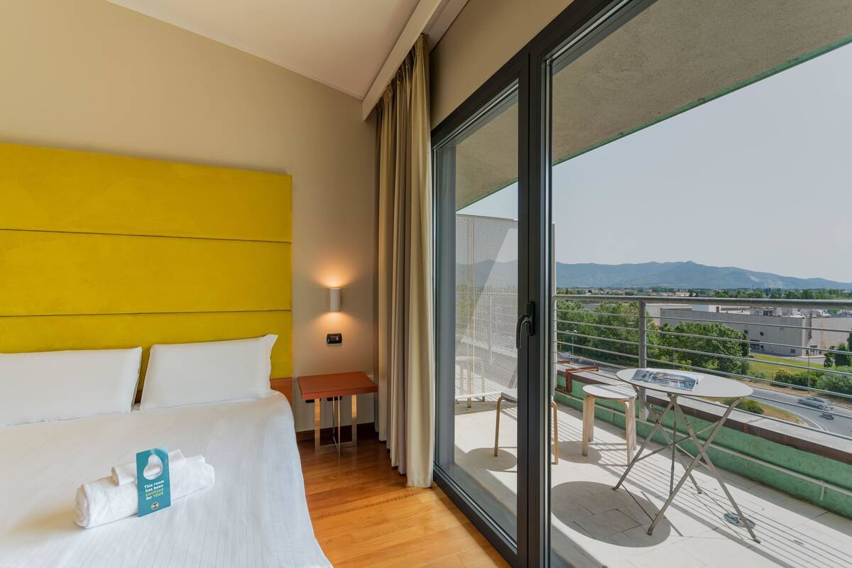 A hotel room with a bed, a small table, a chair, and a balcony with a view of the city.