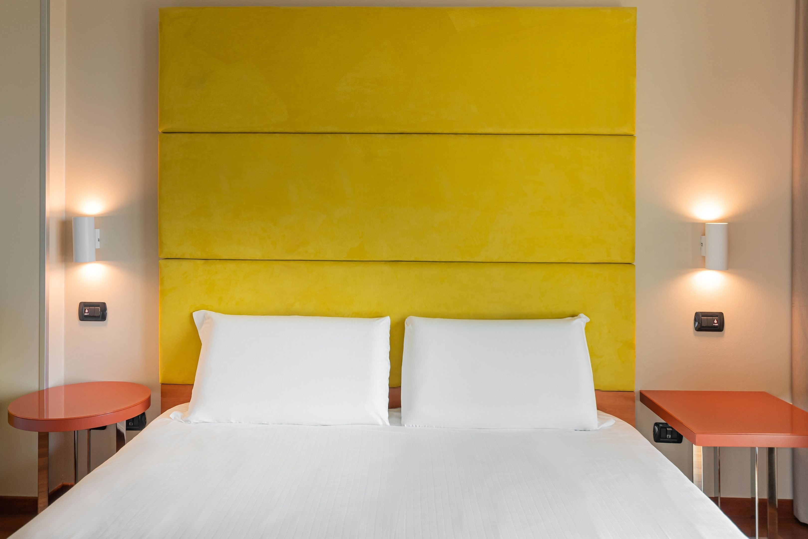 A hotel room with a large bed, two bedside tables, and a bright yellow accent wall.