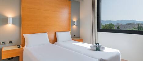 Habitación de hotel con dos camas, cabecero de madera, lámparas de noche, una mesita de noche, un ventanal con vistas a edificios y montañas, y una pequeña mesa con un libro y una toalla.