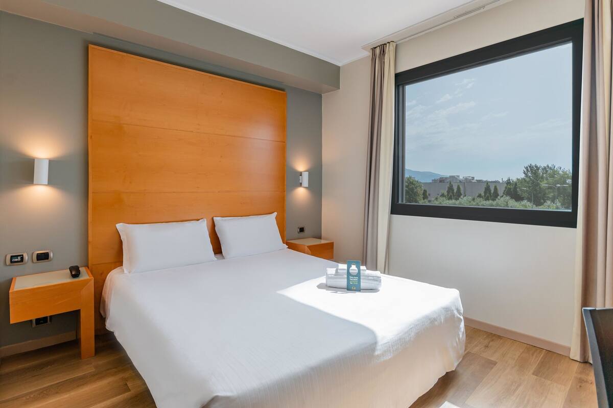 A modern hotel room with a large bed, wooden headboard, bedside lamps, a nightstand, and a view of the cityscape through a large window.
