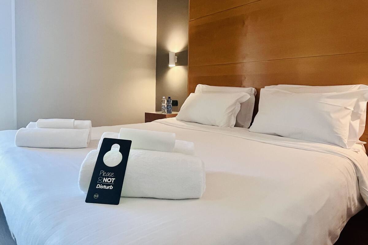 A neatly made bed with white linens and pillows, a bedside table with a water bottle, and a sign that reads "Please DO NOT Disturb".