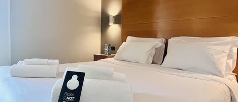 A neatly made bed with white linens and pillows, a bedside table with a water bottle, and a sign that reads "Please DO NOT Disturb".